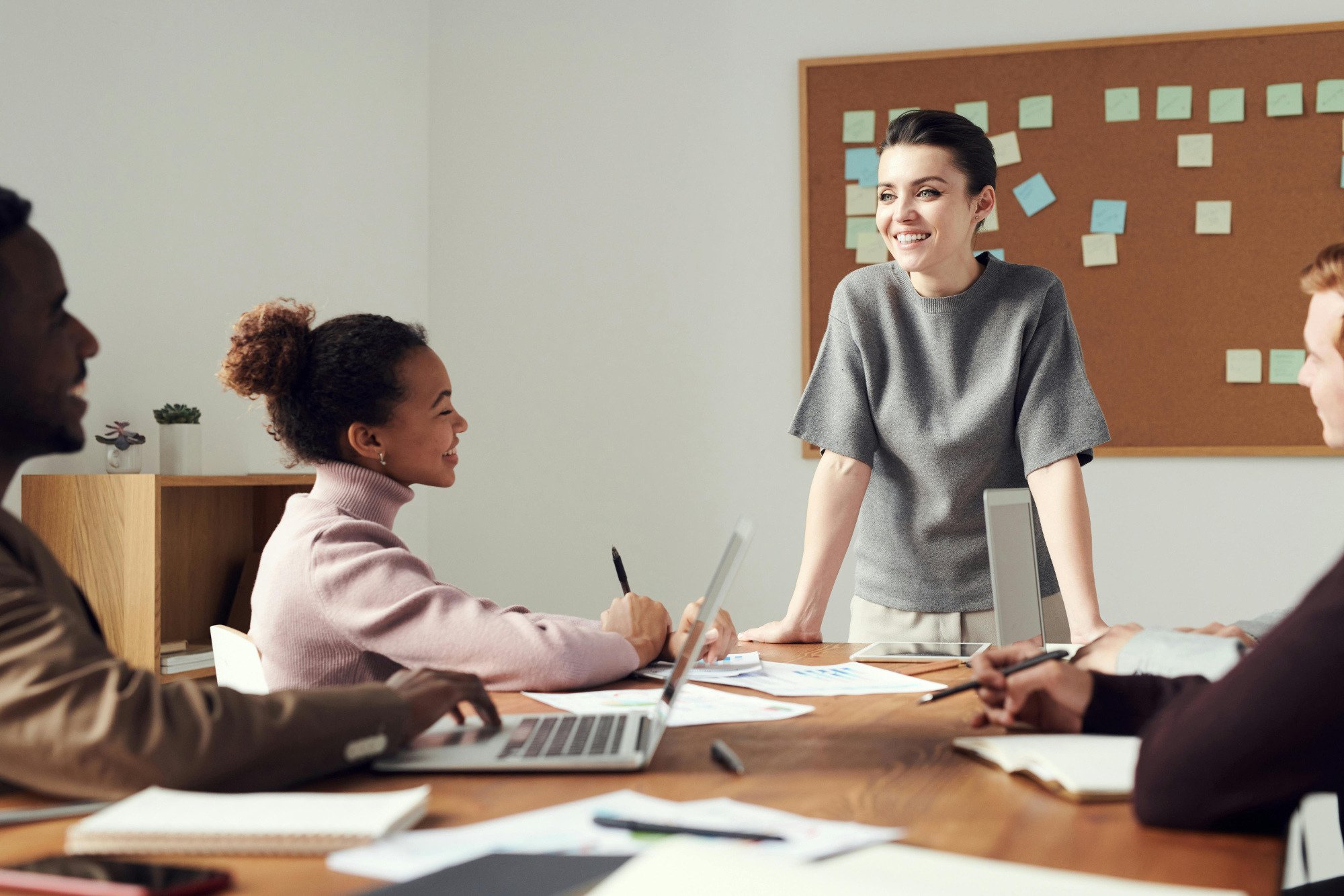 Three people sitting around desk with another person at the end of the desk looking down at them.