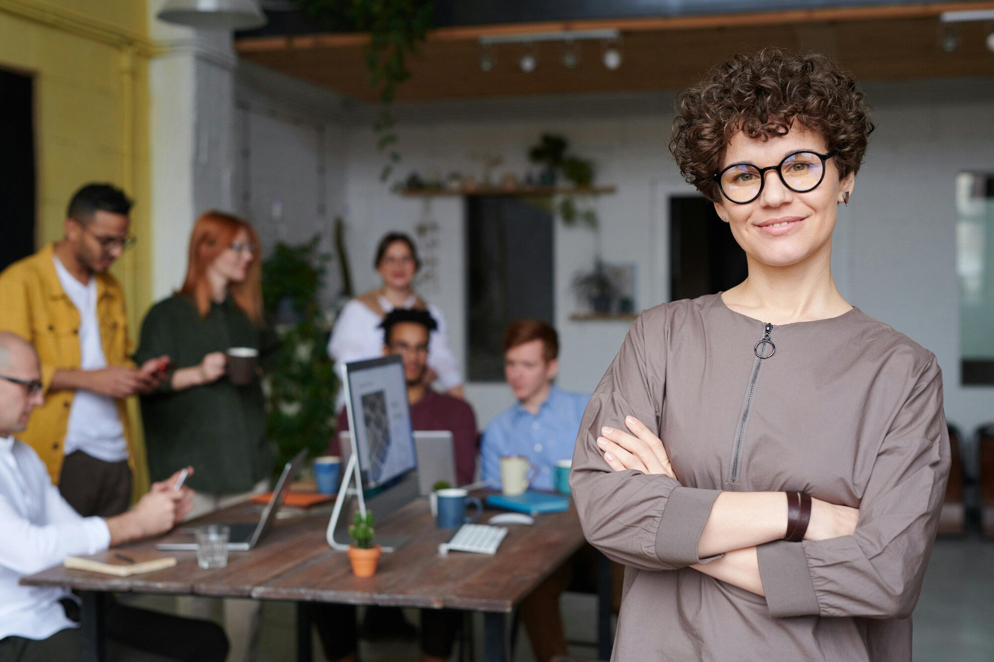 Office worker standing with arms folded with other office workers working at table behind her.