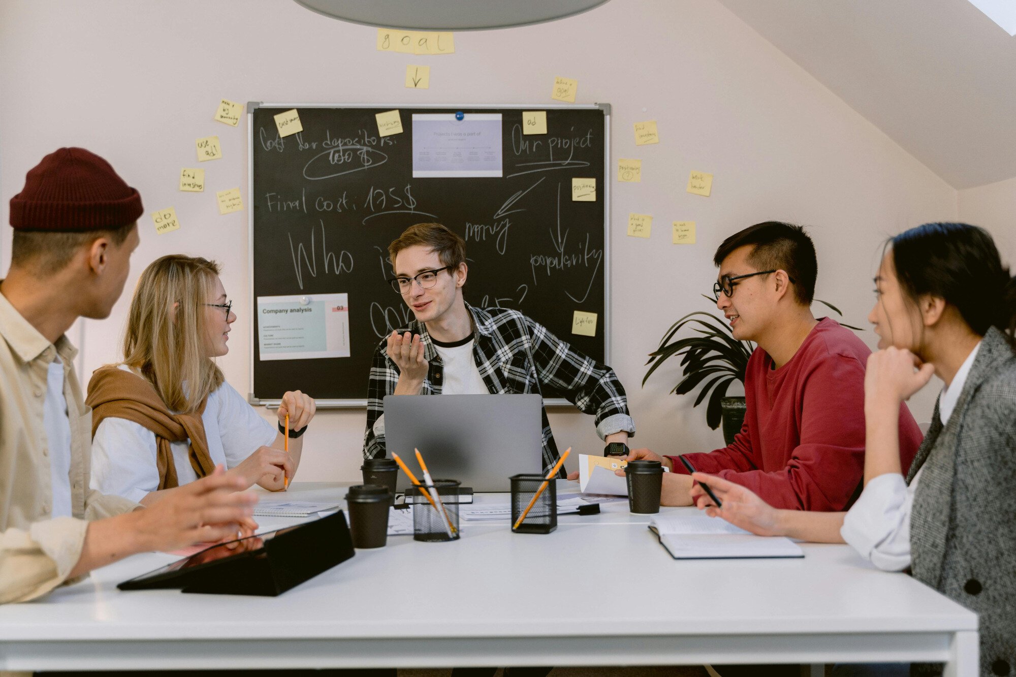 Five office workers sitting around desk with blackboard behind them.