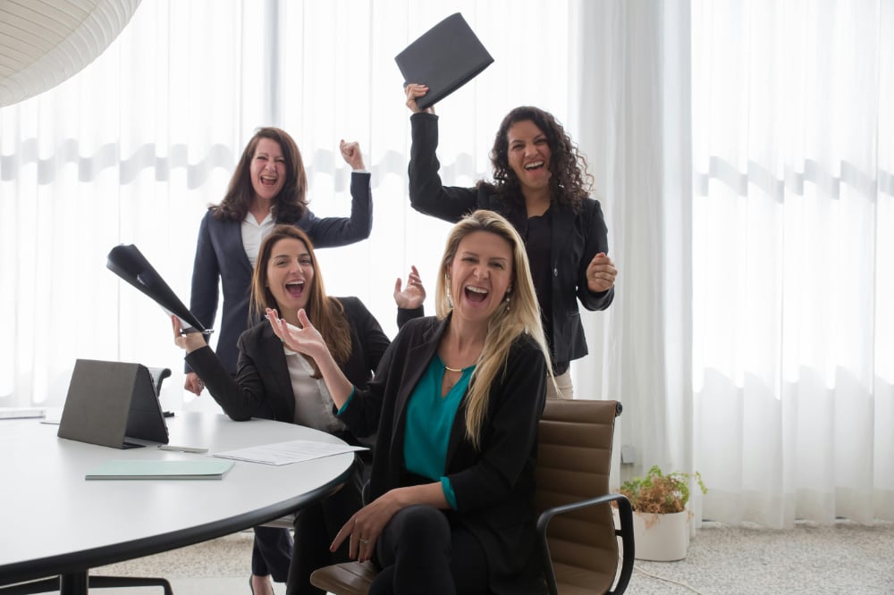 Four office workers holding up folders and celebrating next to desk.