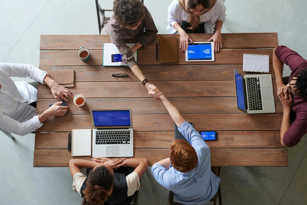 Business people sat around wooden table with two reaching across table and shaking each other's hands.