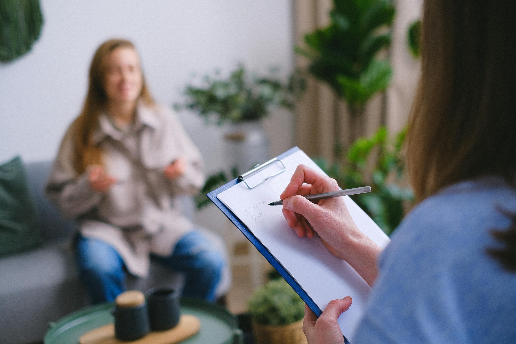 Woman writes on a notepad while sitting across from another woman sitting on a couch.