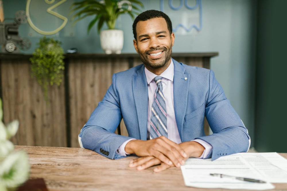 Interview candidate in blue suit smiling with hands resting on wooden desk.