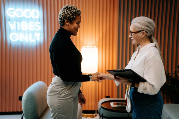 Two women standing in front of desk in office shaking hands.