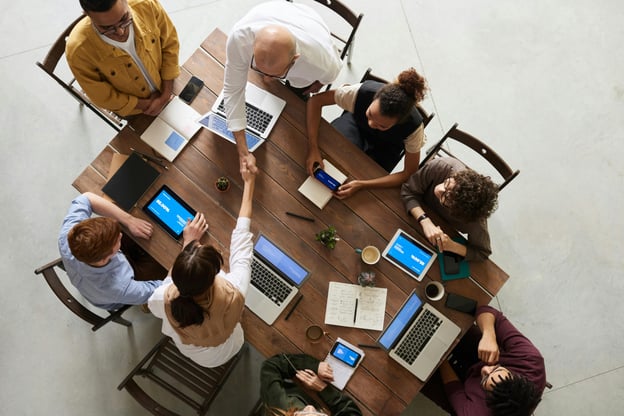 Above view of employees sitting around wooden office table with laptops out.