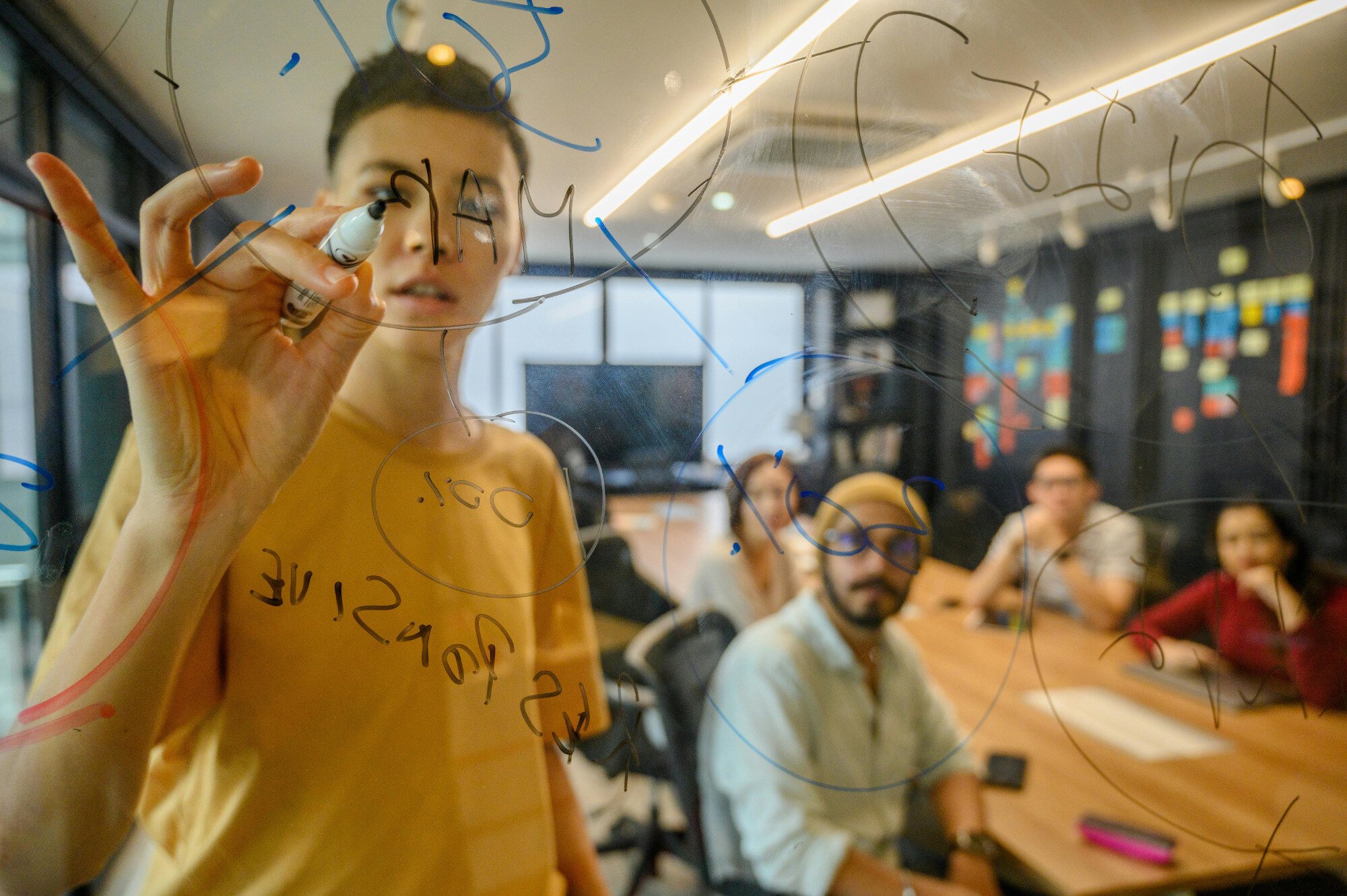 View through a transparent writing board of somebody writing on it with others looking on.