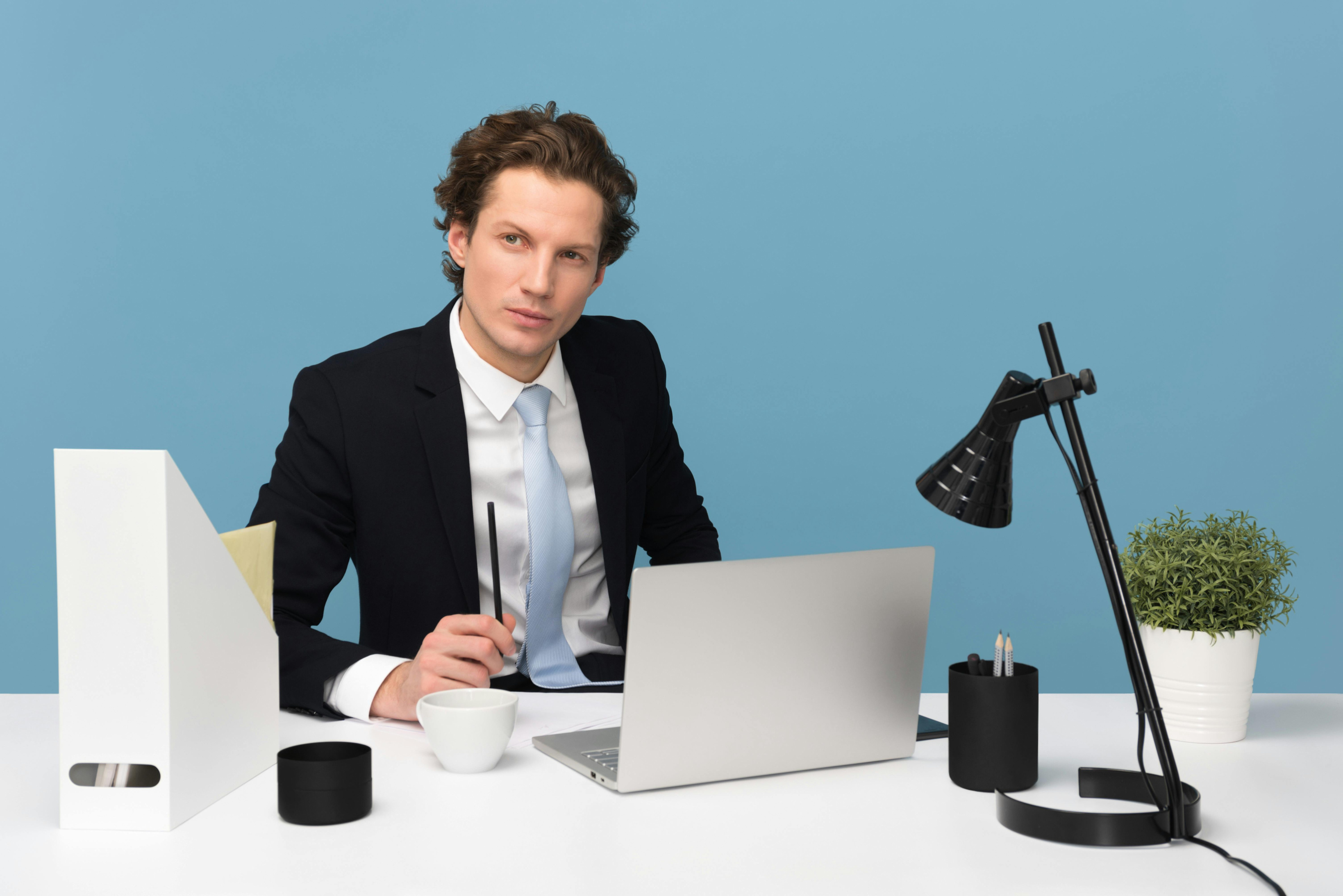 Man sitting at desk in front of blue wall with laptop, lamp, and plant pot on the desk.