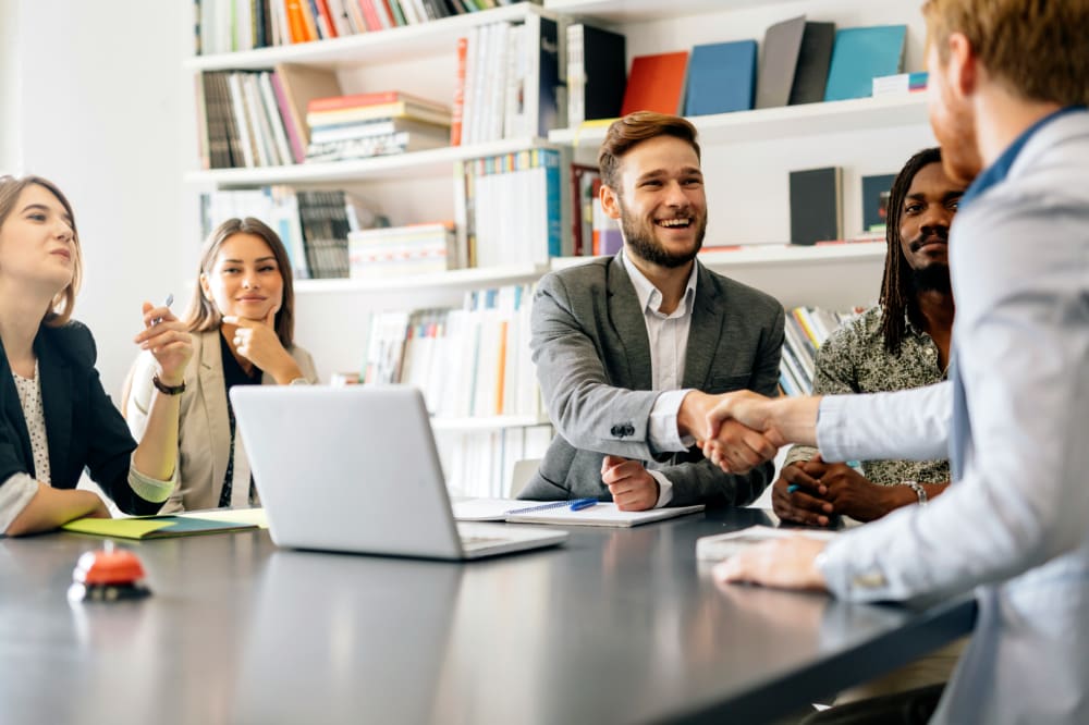 Two office workers shaking hands at desk surrounded by other workers.
