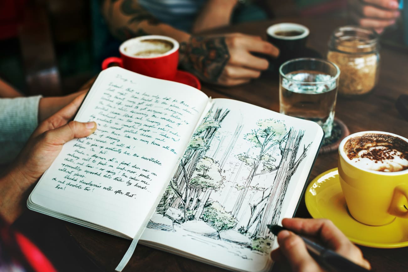 Somebody sitting at a coffee table with an open notepad with sketches of trees in it.