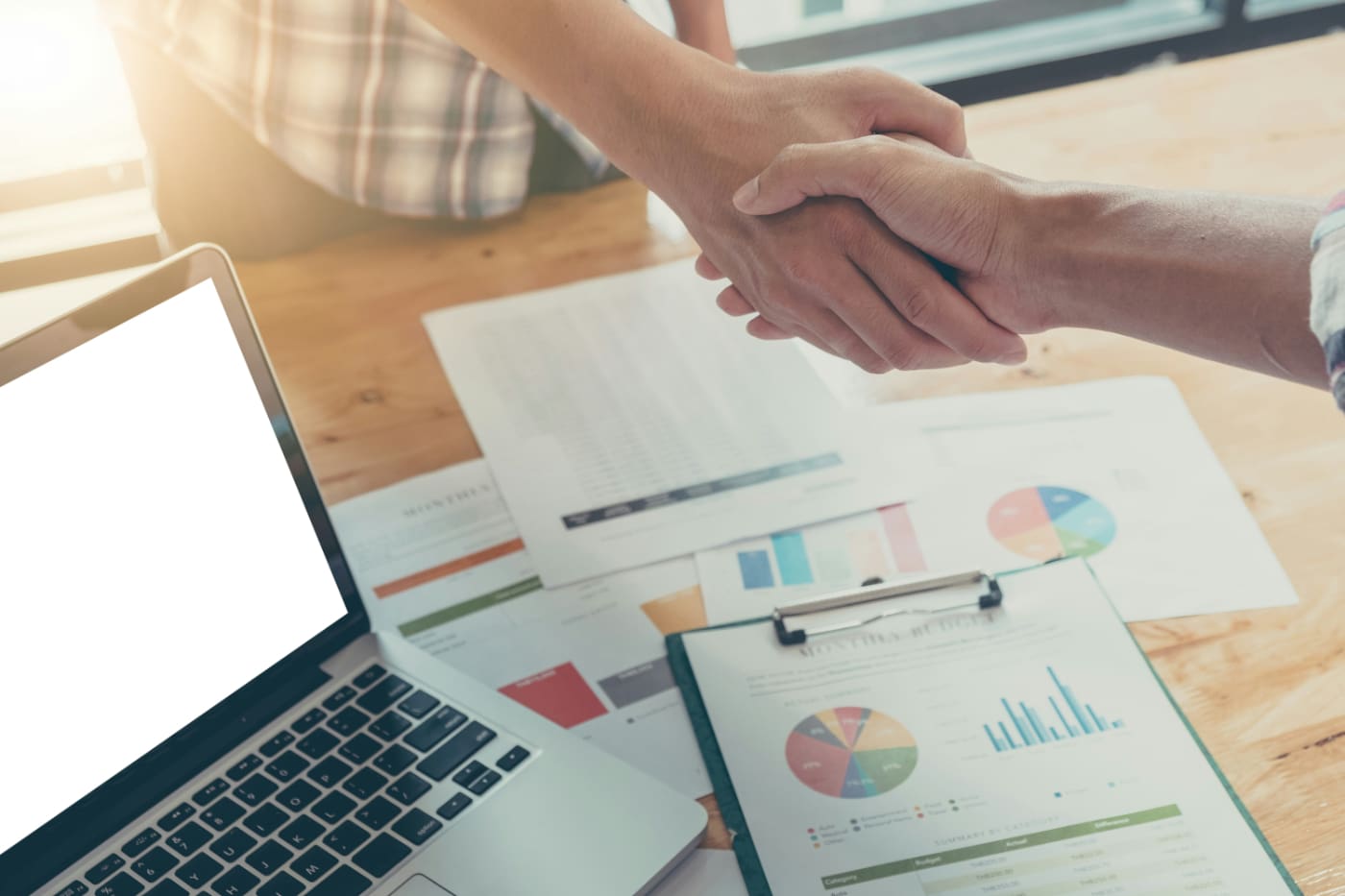 Two business workers shaking hands with business charts sitting on table in the background.