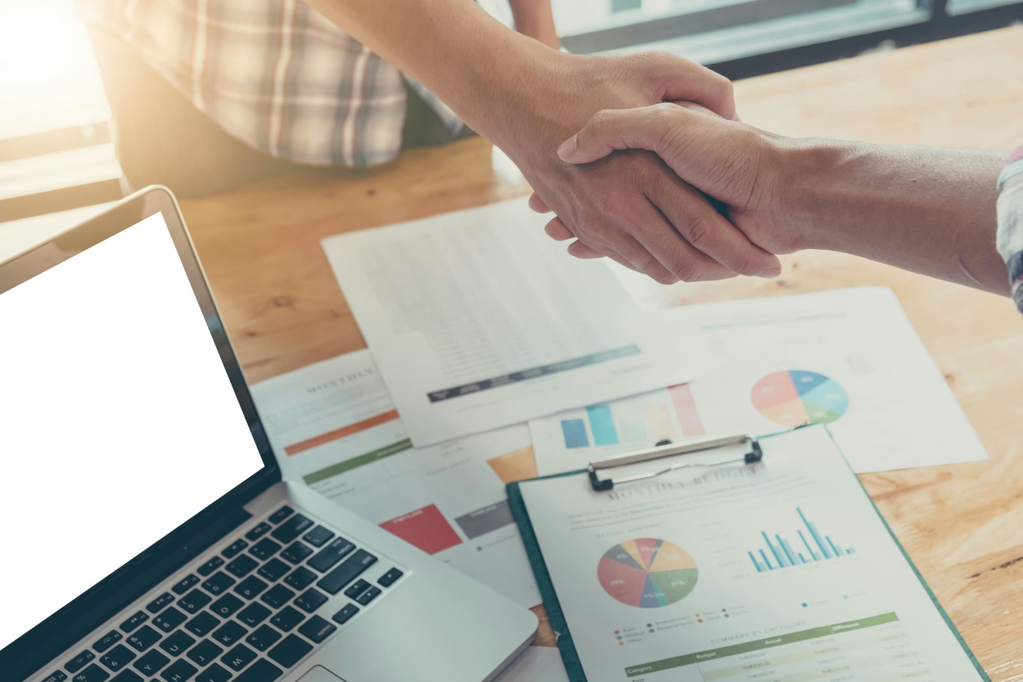 Two business people shaking hands over business charts sitting on desk.