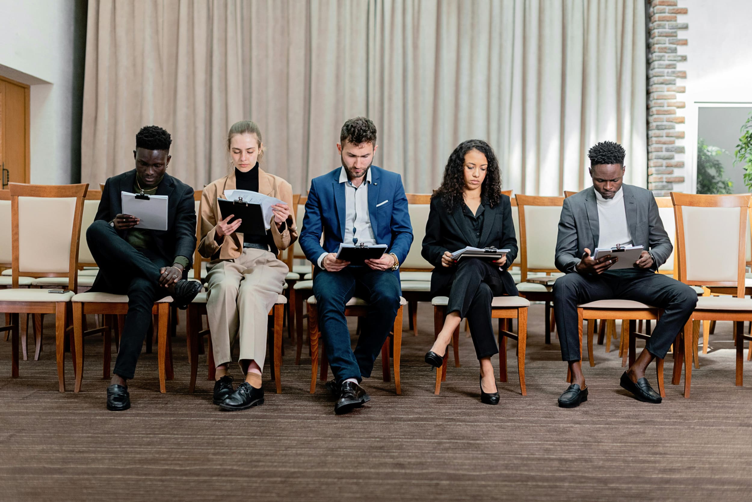 Five interview candidates sitting side-by-side on chairs looking down at their clipboards.