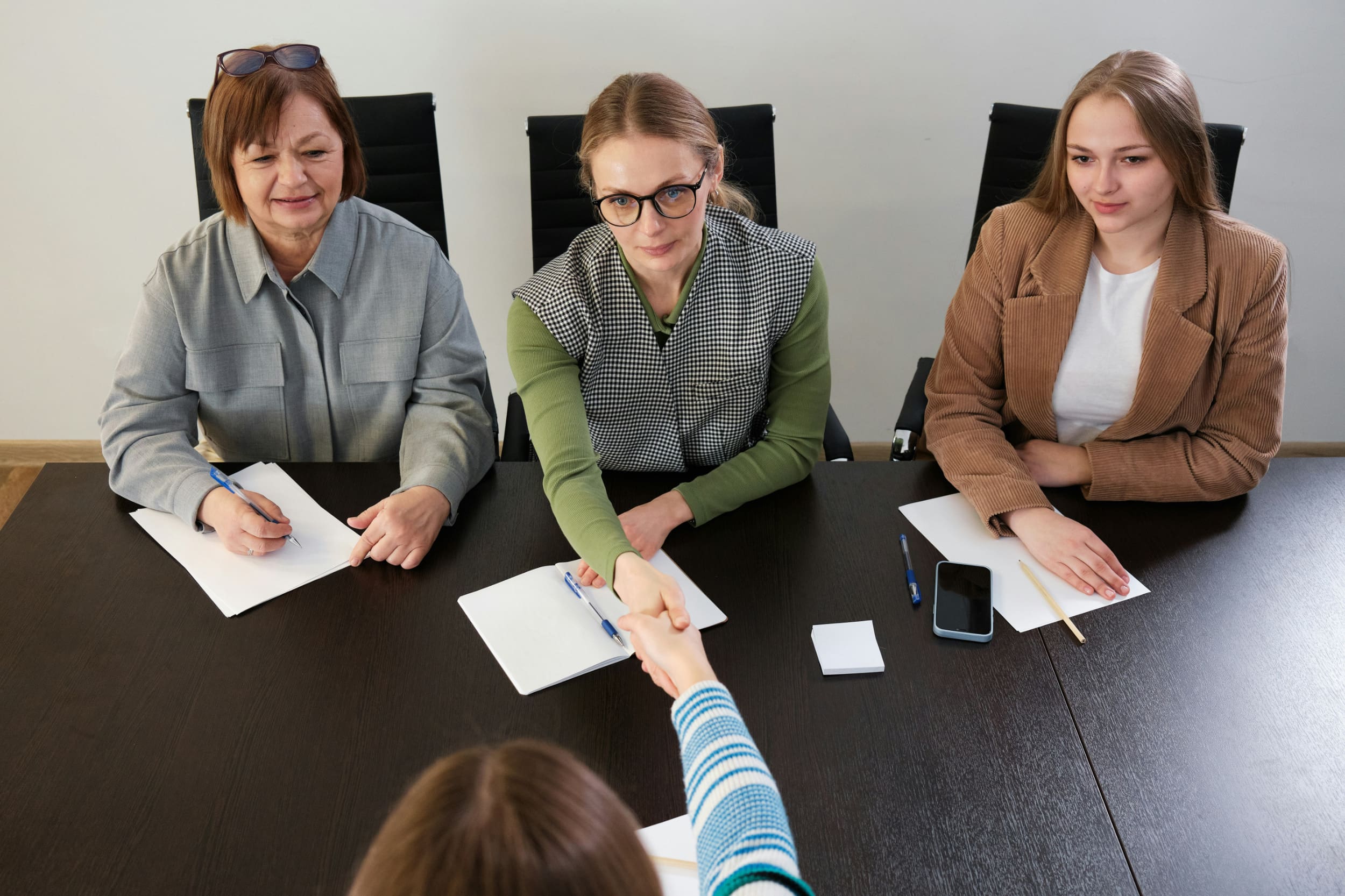 Three women sat on far side of office desk with documents in front of them shaking hand of a woman on other side of the desk.