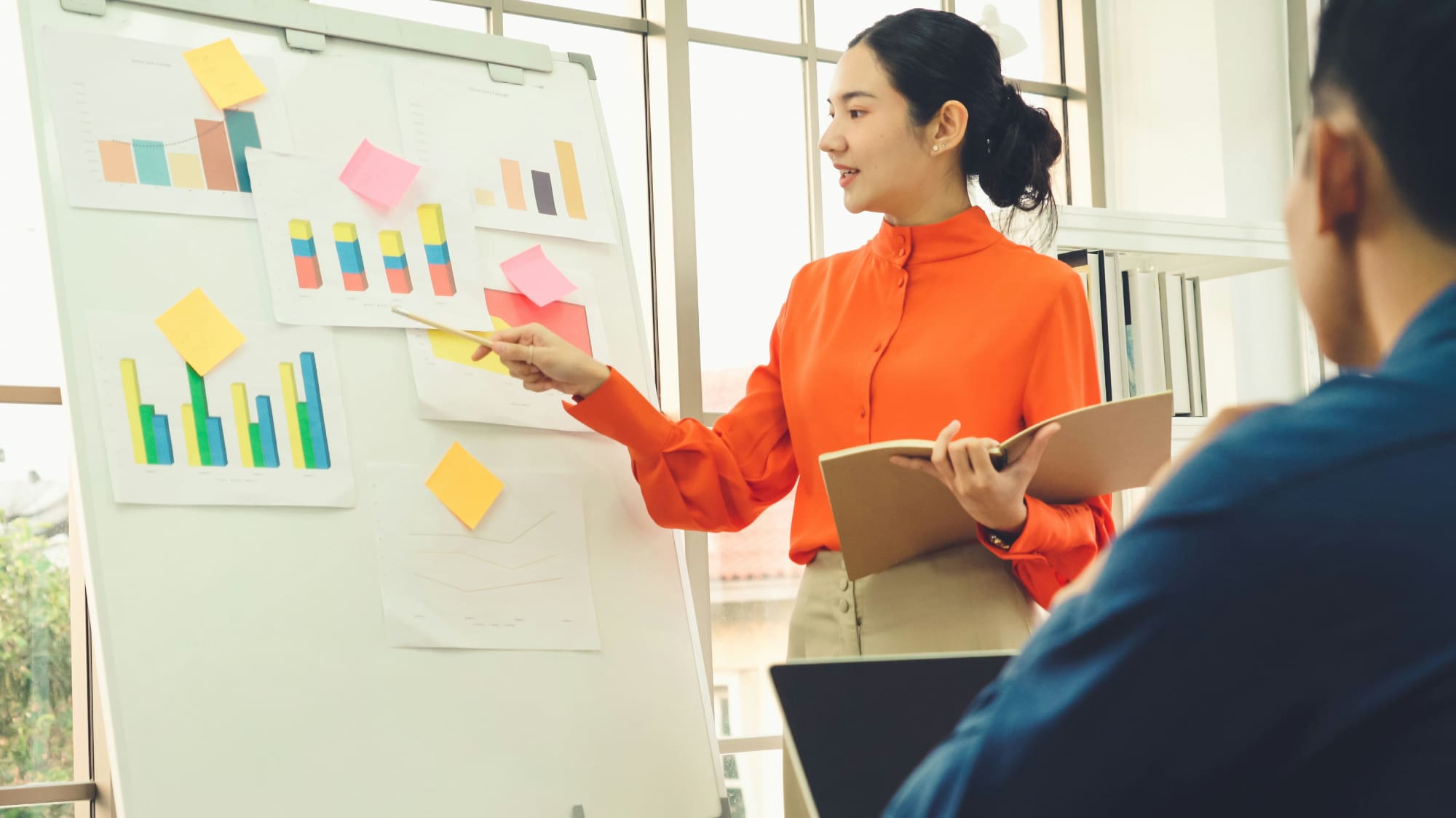 Woman pointing to whiteboard covered in business charts with man looking on.