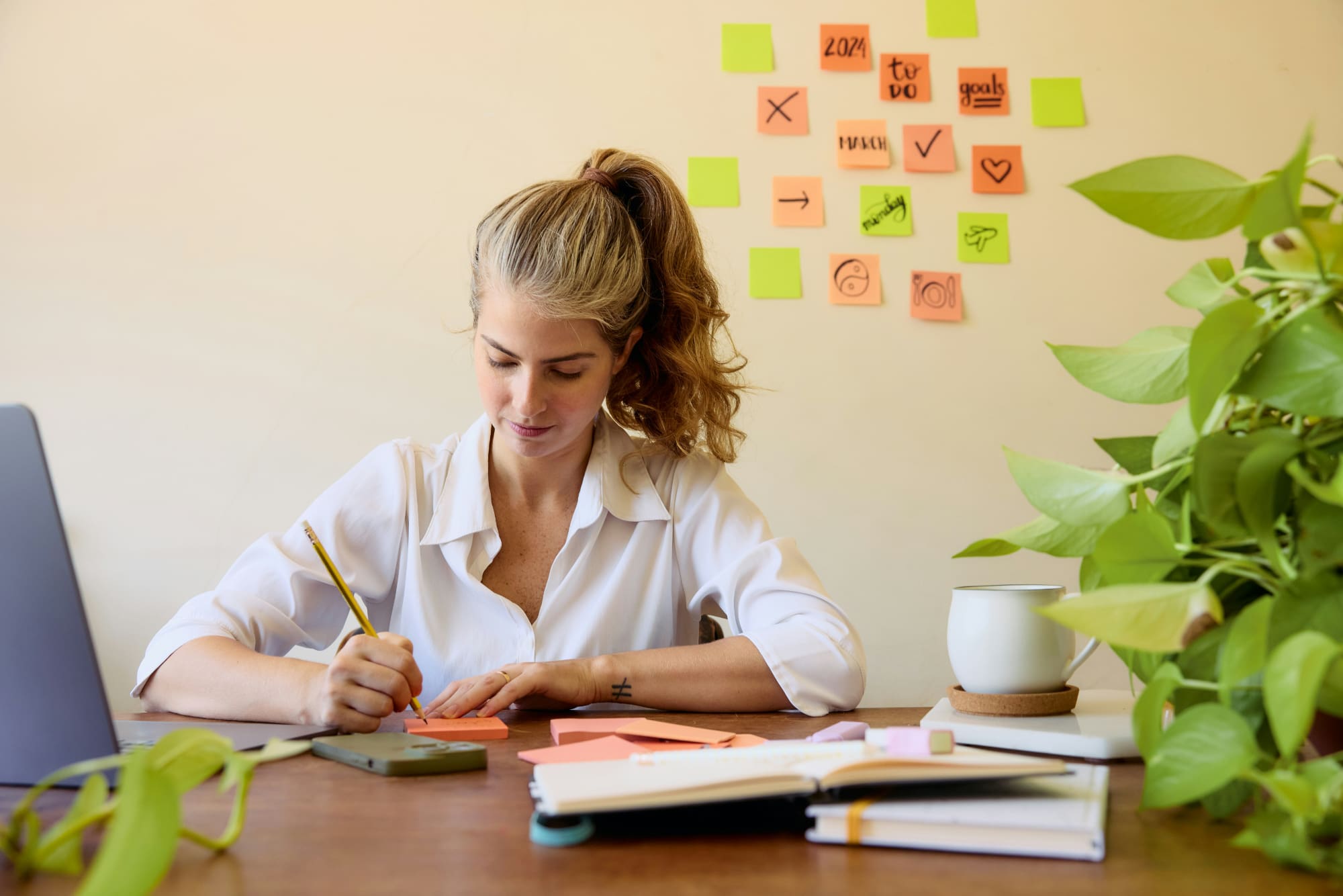 Woman sitting at work desk with plant beside her and post-it notes stuck to wall in the background.