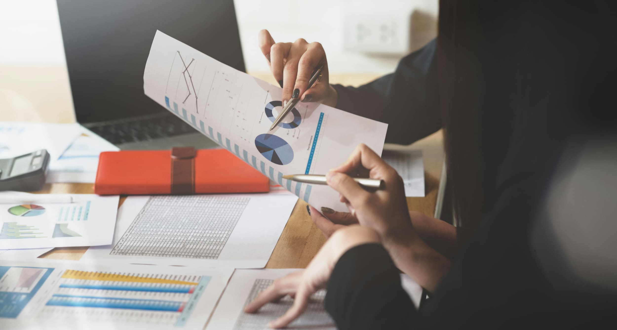 Two business people sat at desk covered in charts and documents holding a document and pointing at it with pens.