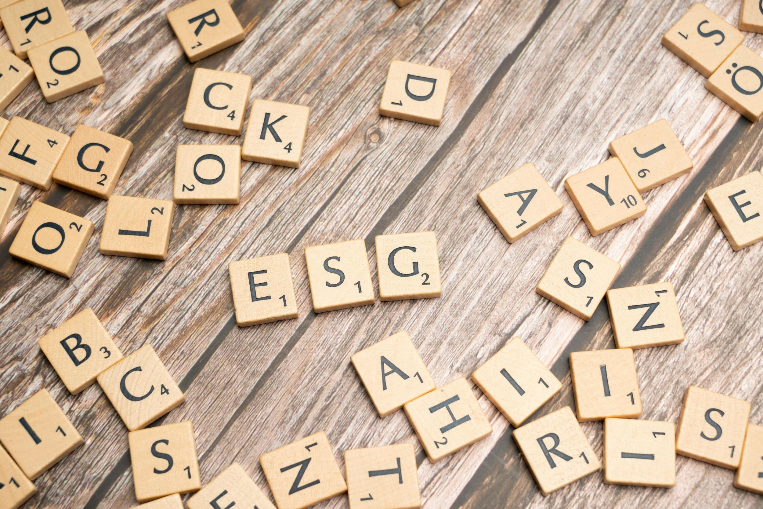 Scrabble pieces scattered on a wooden table with the pieces 'ESG" together in the middle.