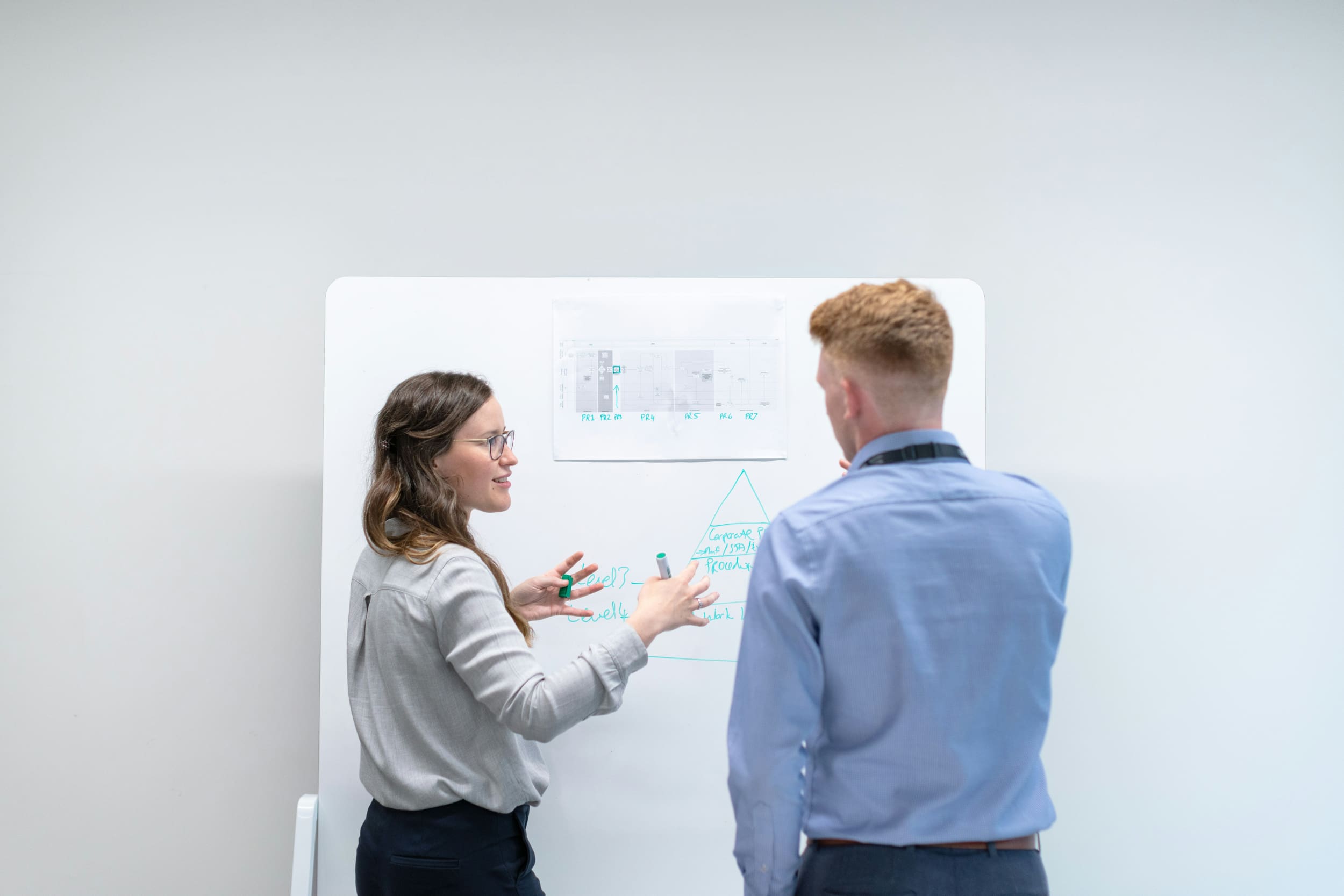 One office worker points to whiteboard to explain something to office worker standing beside her.