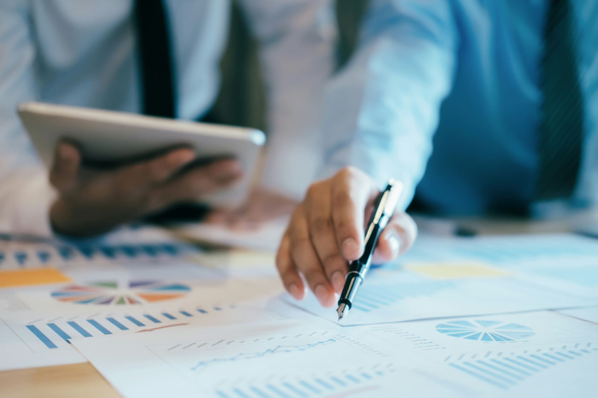 A business person pointing at a printed chart while another one looks at a tablet.