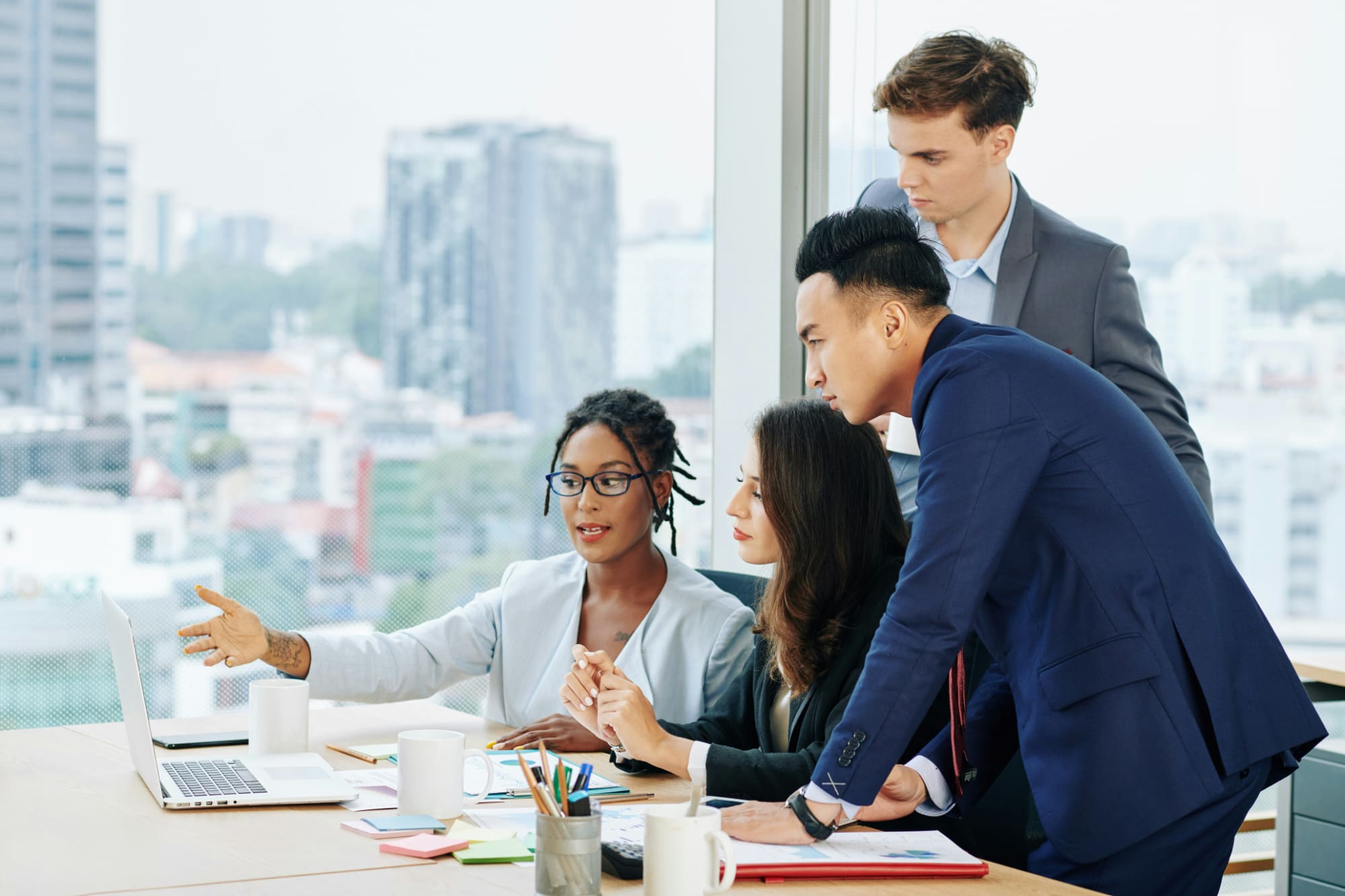 One business person pointing at laptop screen on desk with three others standing behind them looking at the screen with big office window in the background looking out onto a cityscape.