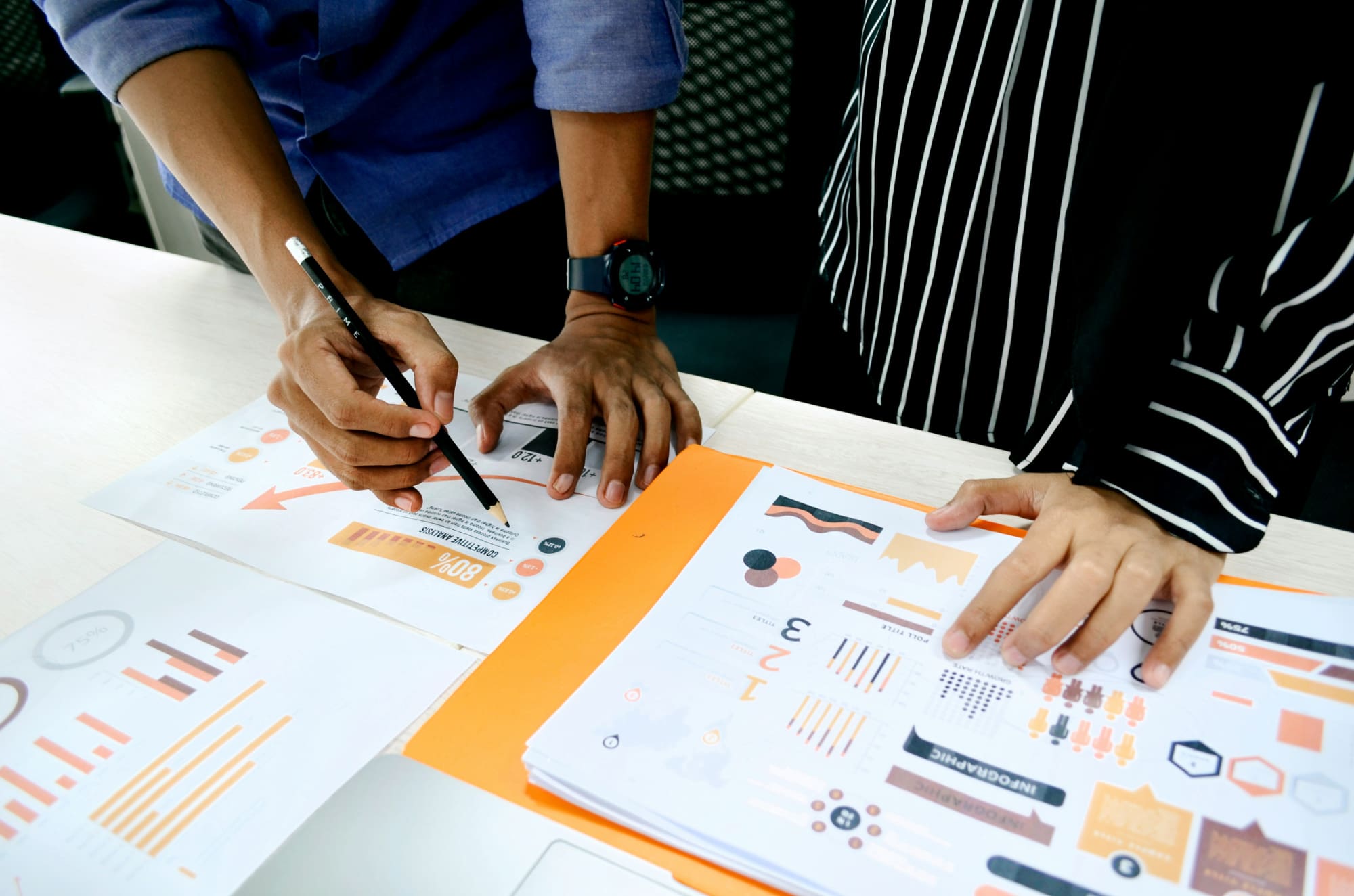 Two people stood behind desk pointing at various paper business documents arranged on the desk.