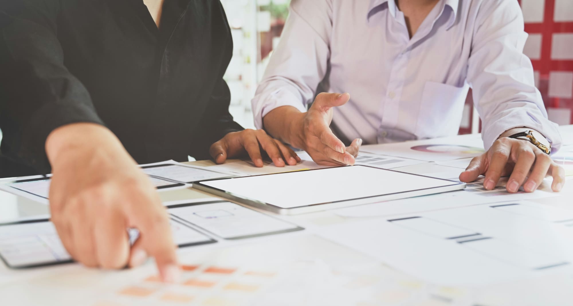 Two people sat behind desk covered in paper documents with one person pointing out something to the other.