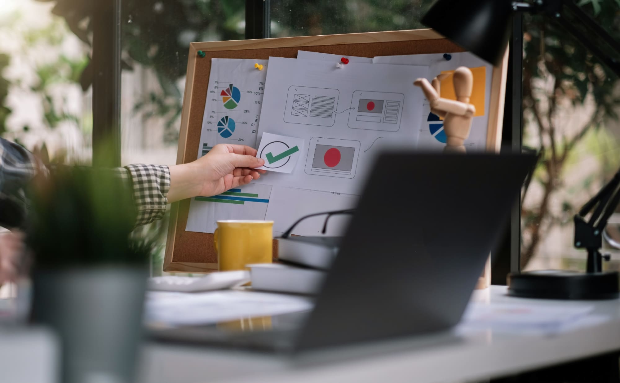 Person placing 'ticked box' sticker onto a message board with laptop on desk in the foreground