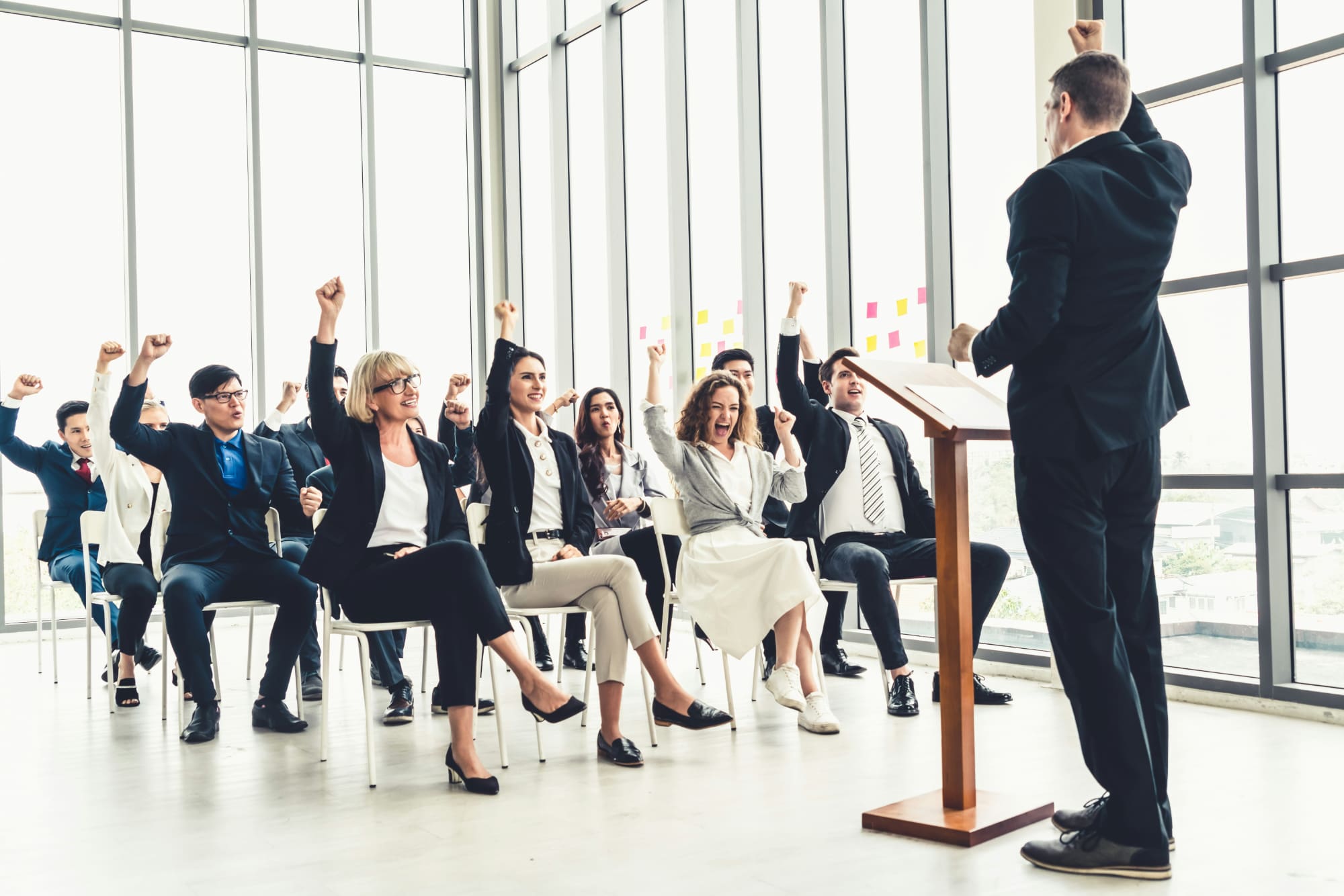 Standing business person giving presentation to other sitting business people who are raising their hands.