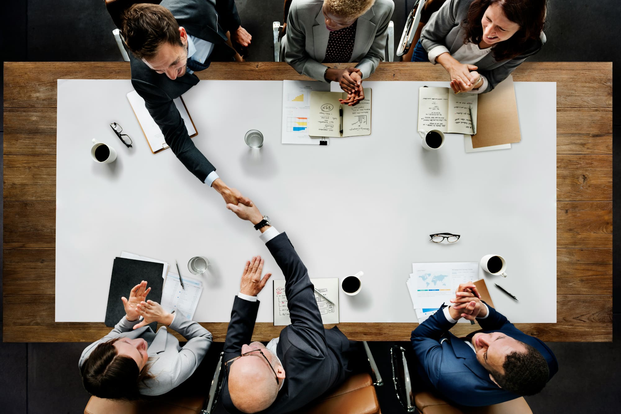 Overhead shot of three business people sitting across table from three others, with two shaking hands across table with notepads, pens, and coffee cups on it.